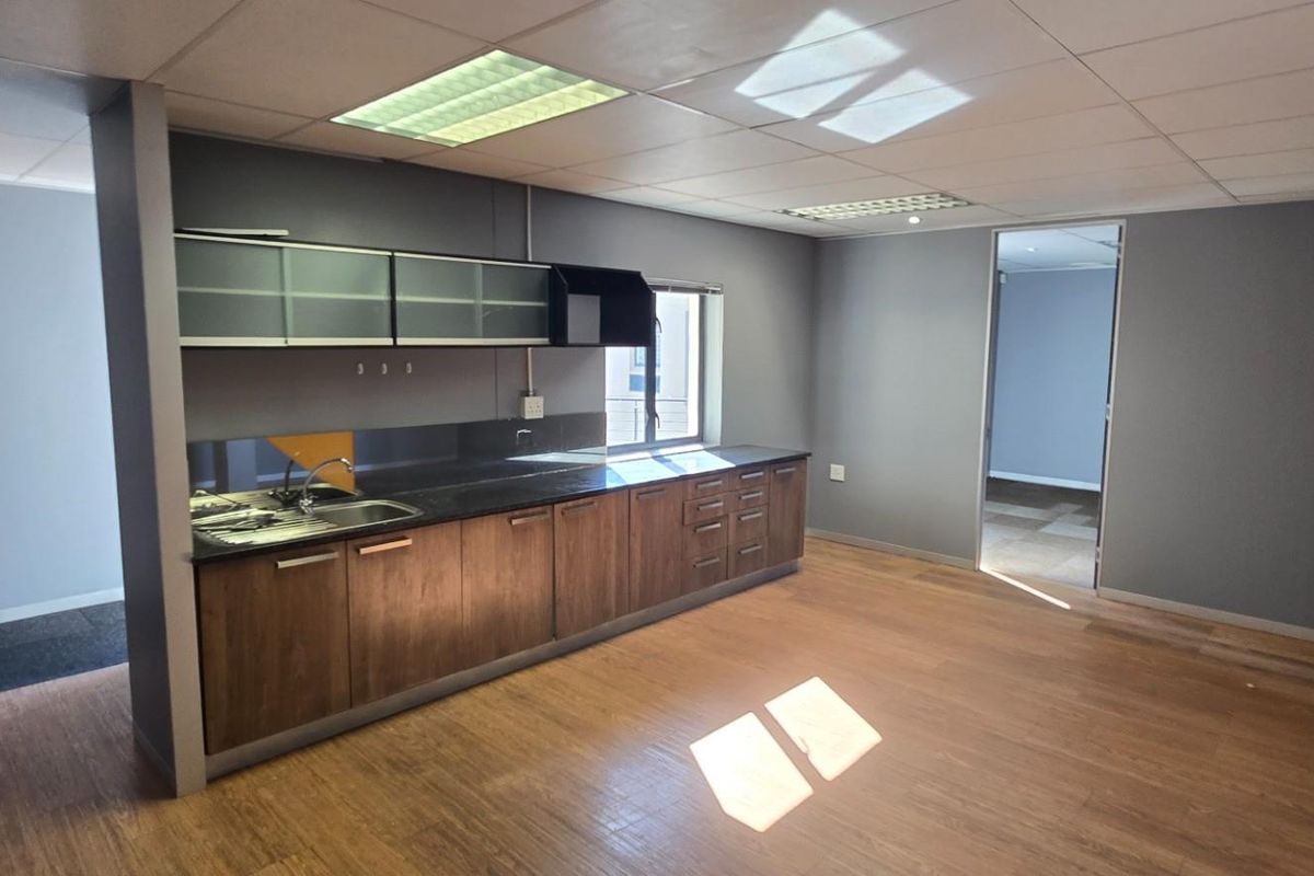 A modern commercial kitchenette featuring wood-grain cabinetry, dark countertops, a stainless steel sink, and integrated overhead storage, set within an open-plan office space.