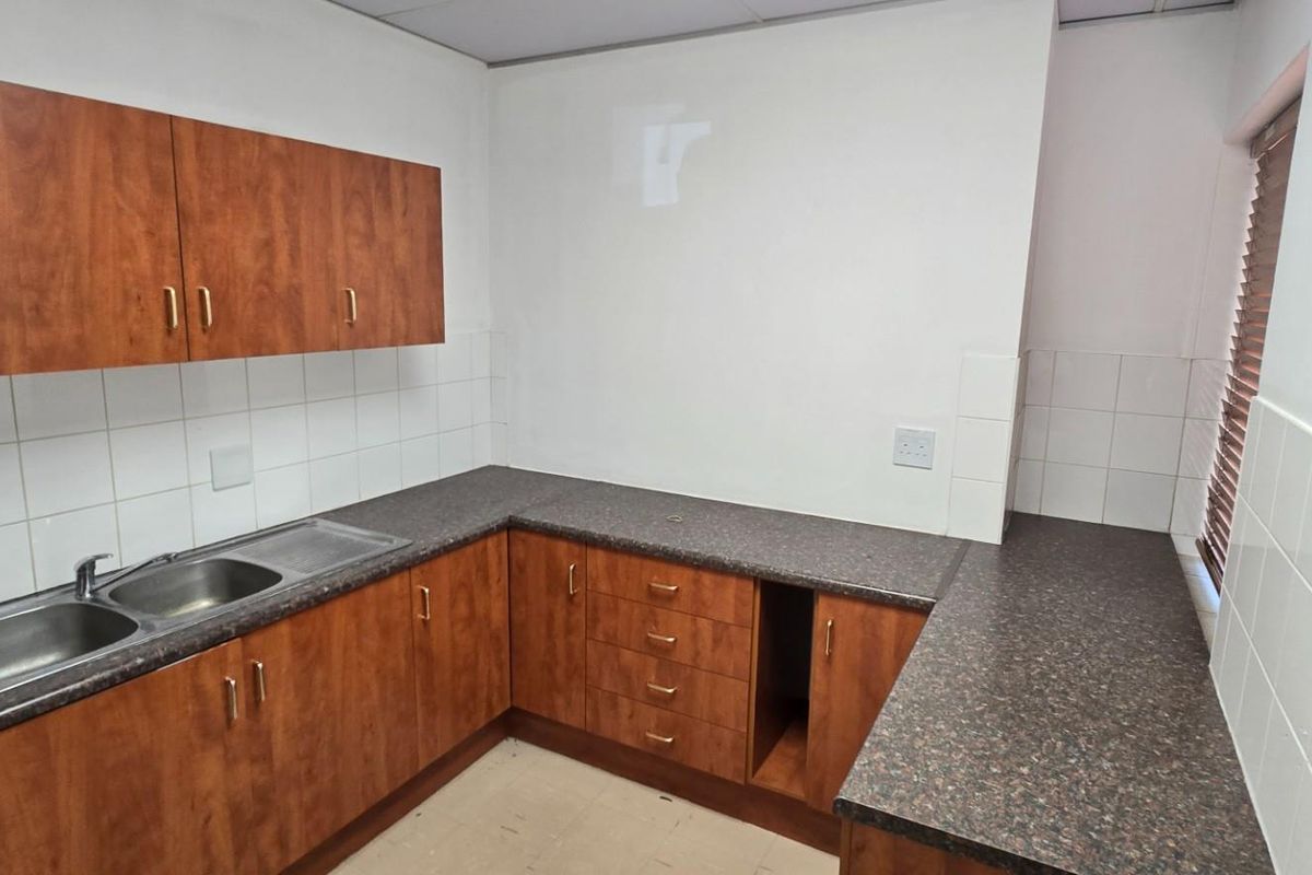 A functional commercial kitchenette featuring wood-finish cabinetry, laminate countertops, a double stainless steel sink, and tiled splashbacks.