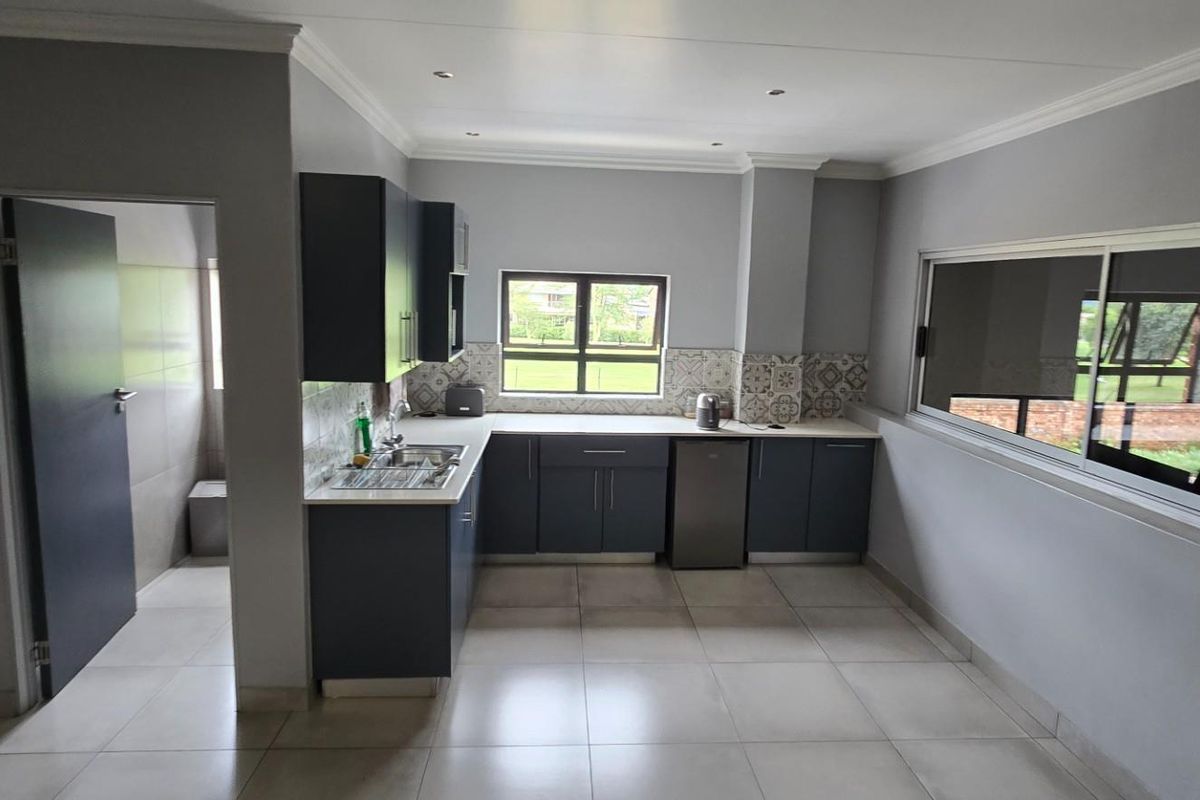 Modern kitchen featuring dark cabinetry, tiled backsplash, and neutral floor tiles, with an adjacent utility area and large windows providing natural light.