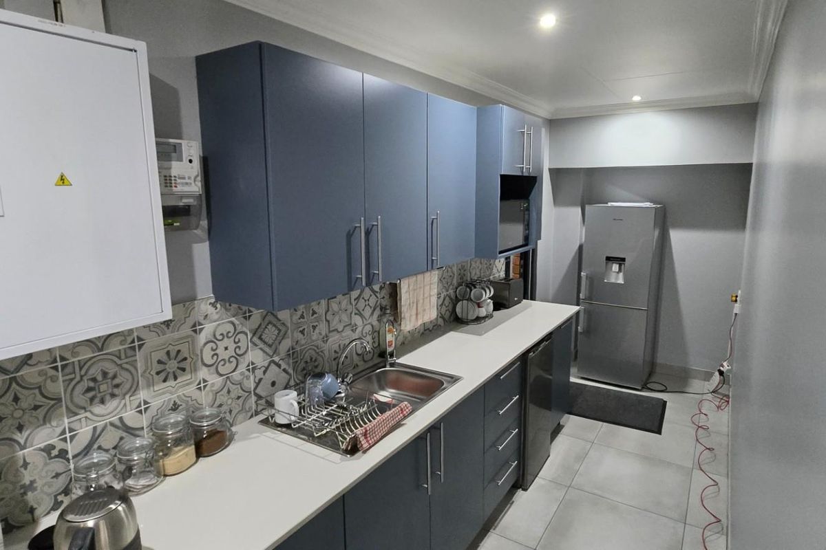 A modern, compact kitchen featuring blue cabinetry, patterned tile backsplash, stainless steel sink, and tiled flooring.