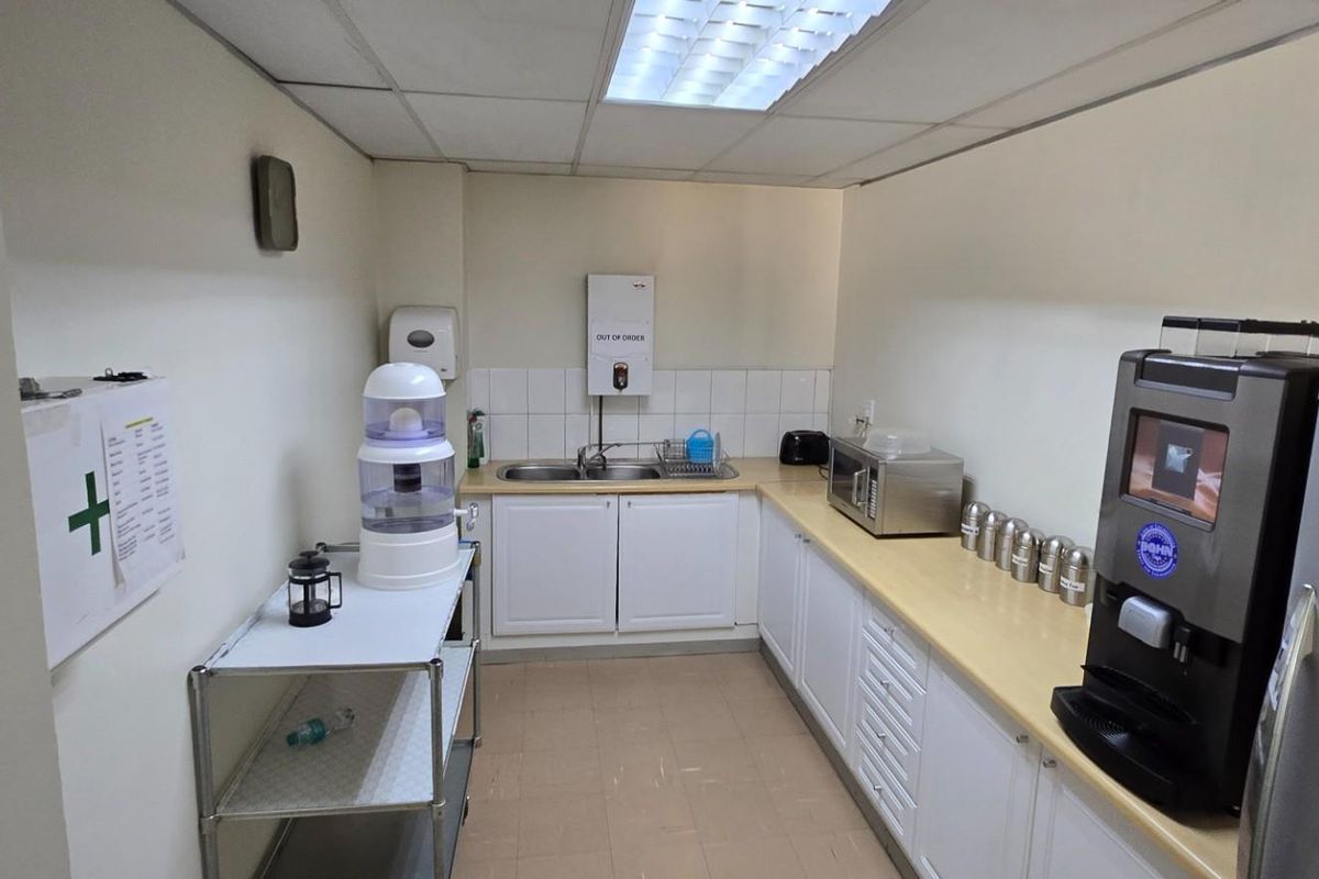 A functional commercial kitchenette featuring built-in cabinetry, a stainless steel sink, tiled splashback, and a suspended ceiling with integrated lighting.