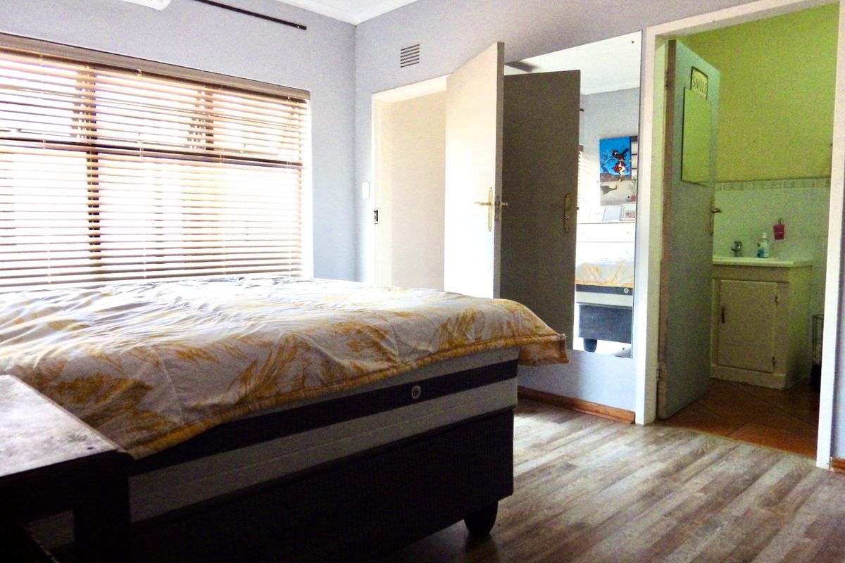 A bedroom featuring wood-look flooring, venetian blinds, and an integrated air conditioning unit, with an adjoining en-suite bathroom visible through the doorway.