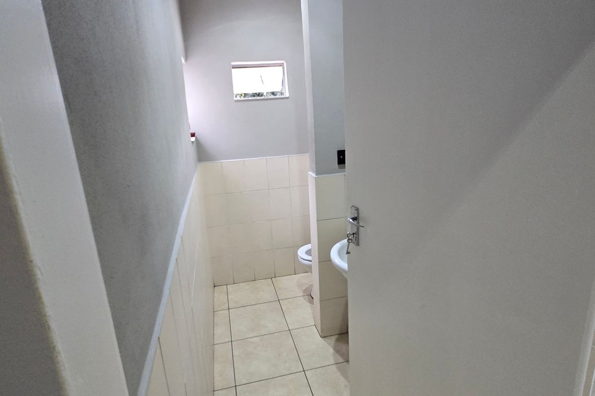 A functional commercial bathroom featuring tiled walls and flooring, a pedestal basin, and a toilet, with a small window for natural light.