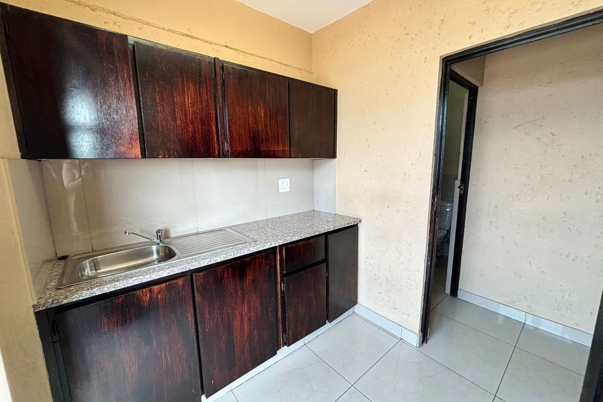 A compact kitchenette featuring dark wood-finish cabinetry, a stainless steel sink, and tiled flooring, with an adjacent doorway leading to a bathroom.