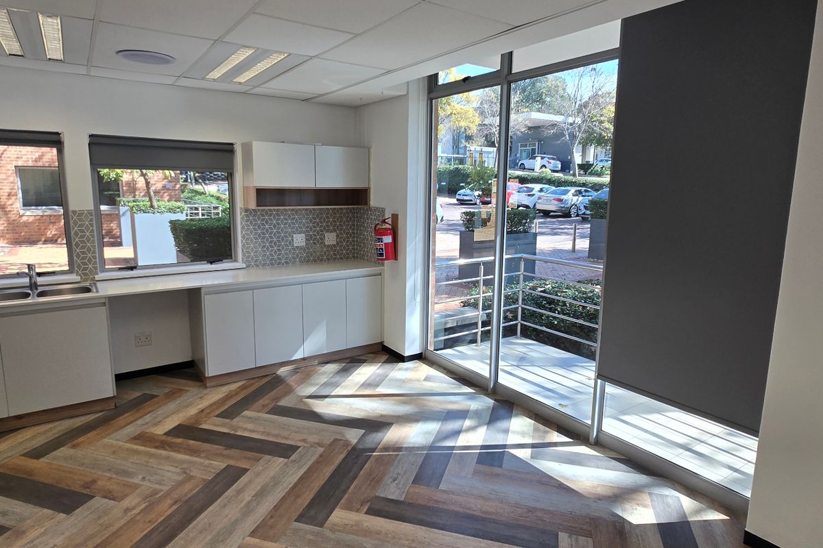A bright, modern office kitchenette featuring herringbone vinyl flooring, built-in cabinetry, a stainless steel sink, and large windows providing natural light and exterior views.