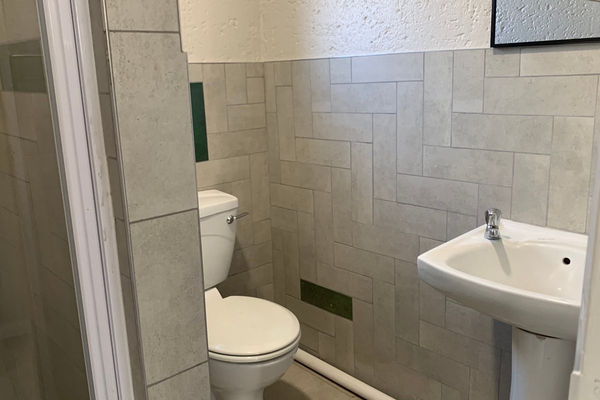 A compact bathroom featuring a pedestal basin, toilet, and shower enclosure with neutral tiled finishes, requiring minor repairs to wall tiling and exposed plumbing.