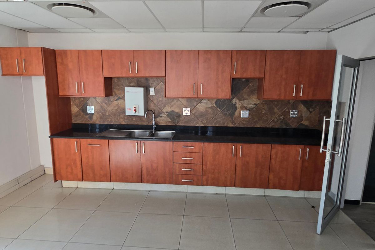 A functional commercial kitchenette featuring wood-finish cabinetry, dark granite countertops, a stainless steel sink, and tiled splashbacks with a suspended ceiling.