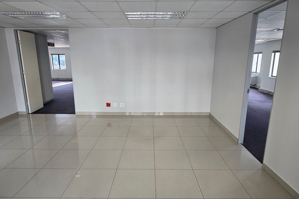 An empty commercial office space featuring tiled flooring, a suspended ceiling with recessed lighting, and multiple doorways leading to adjacent office areas.