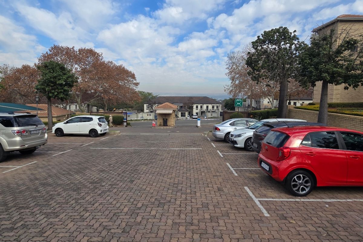 A paved commercial parking area with marked bays, featuring a security access gatehouse in the distance and surrounding landscaped trees.