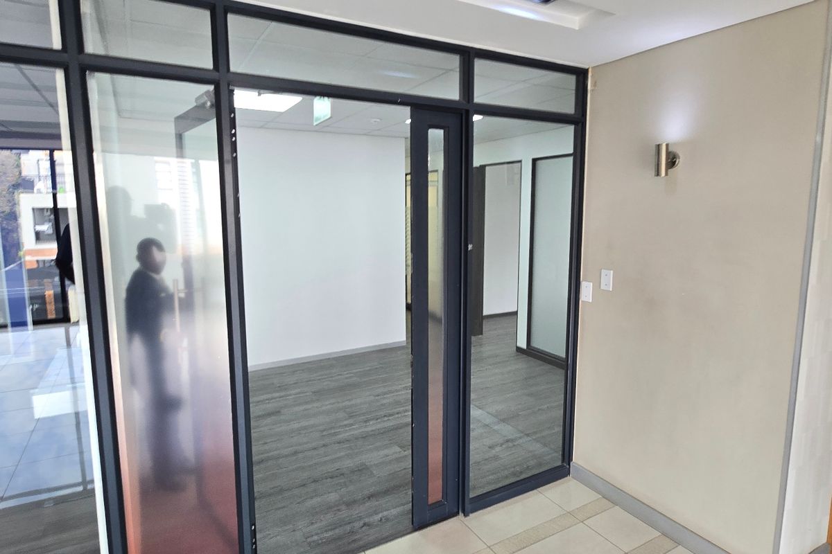 A modern commercial office space featuring glass partition walls, dark wood-look flooring, and recessed ceiling lighting.