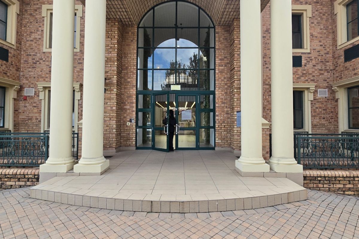 A grand commercial entrance featuring a paved forecourt, prominent structural columns, and a large arched glass doorway providing access to the building interior.