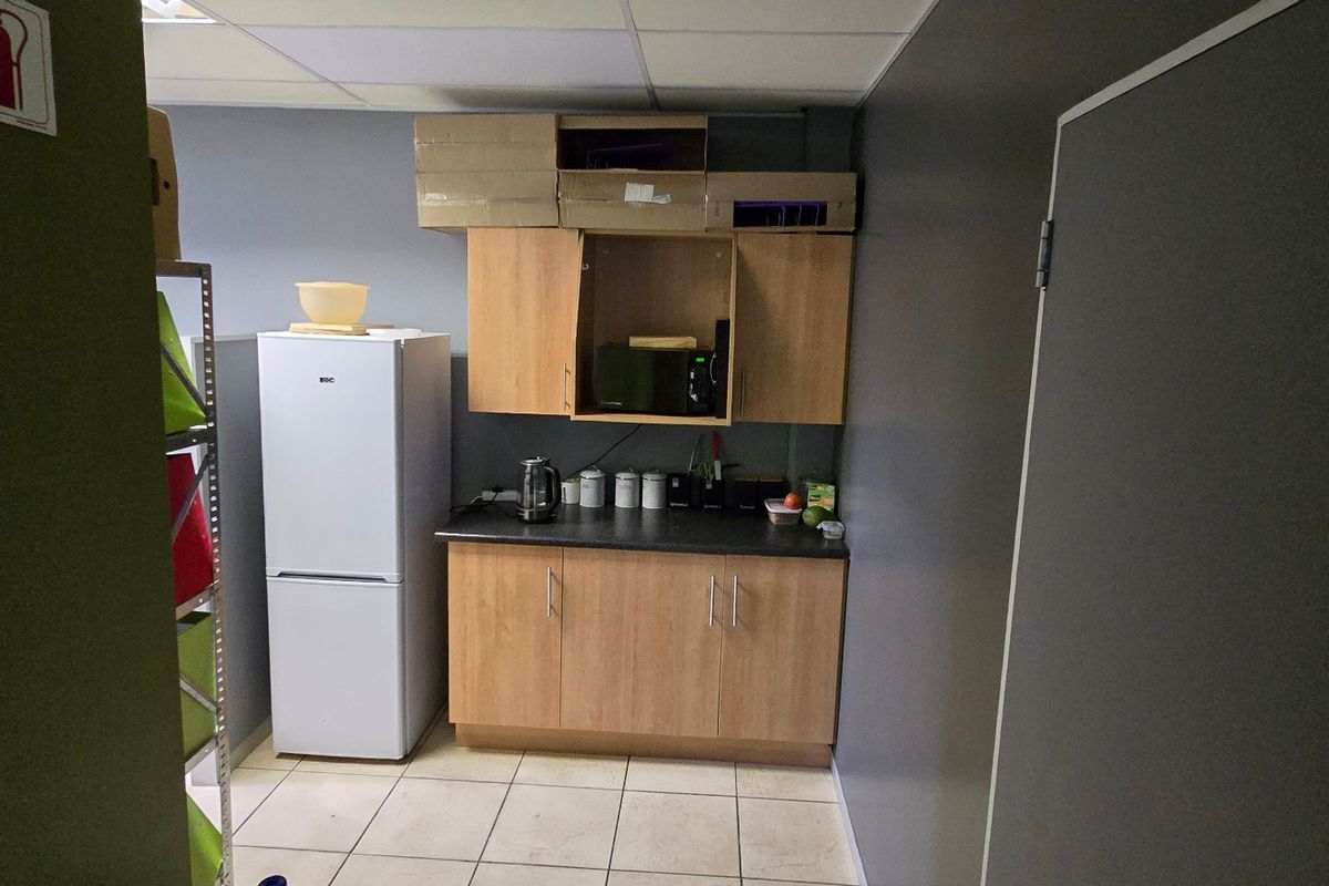 A compact commercial kitchenette featuring light wood-finish cabinetry, dark laminate countertops, and tiled flooring, suitable for office staff use.