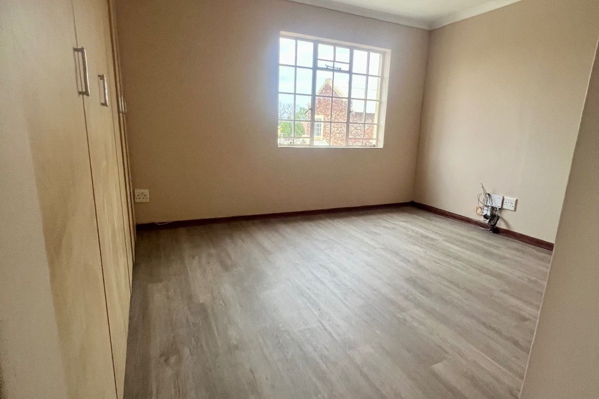 A bright, empty bedroom featuring light wood-look laminate flooring, built-in cupboards, and a large window providing natural light.
