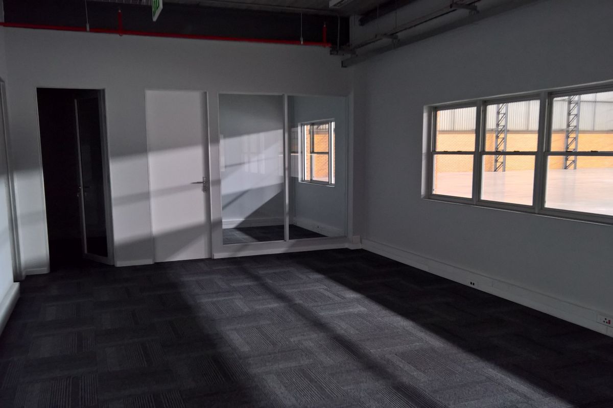 A clean, vacant office space featuring carpet tiles, exposed ceiling services, and large windows providing natural light and views into the industrial warehouse area.