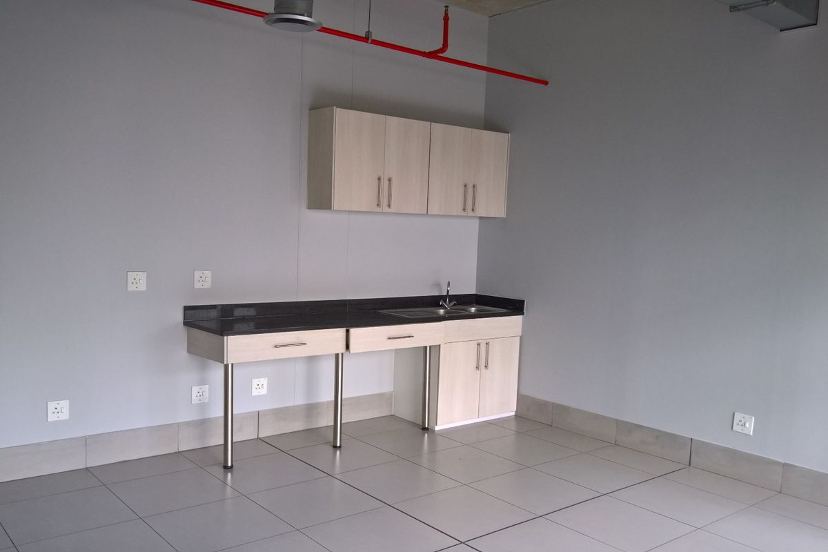 A clean, modern kitchenette area featuring light wood cabinetry, a dark stone countertop, stainless steel sink, and industrial-style exposed fire sprinkler piping.