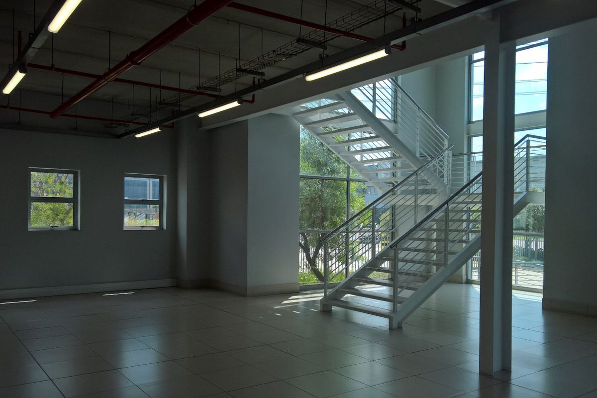 Spacious open-plan industrial office area featuring tiled floors, exposed ceiling services, and a modern metal staircase with large windows providing natural light.
