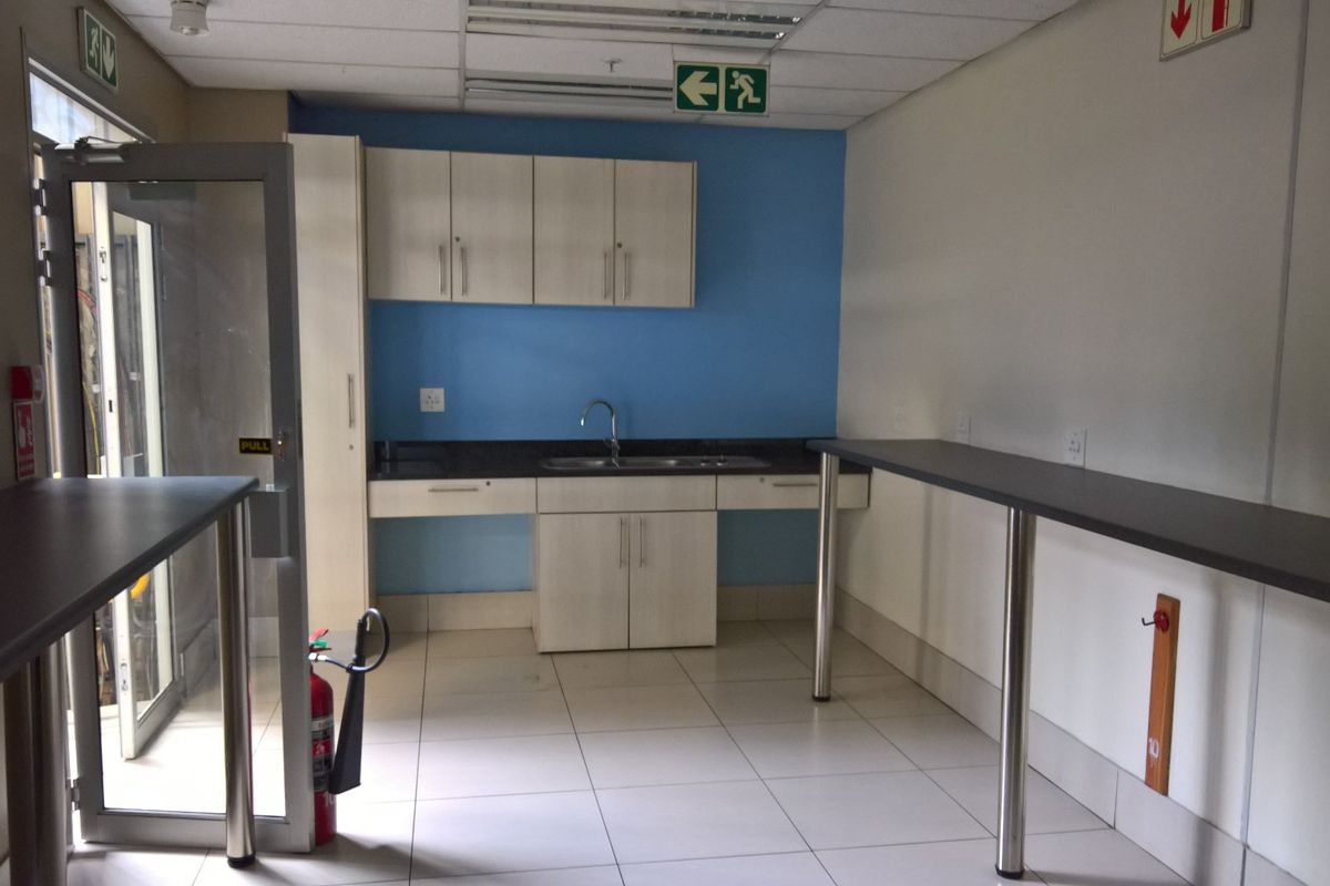 A functional kitchenette area featuring built-in cabinetry, a stainless steel sink, and durable tiled flooring, suitable for office or industrial utility use.