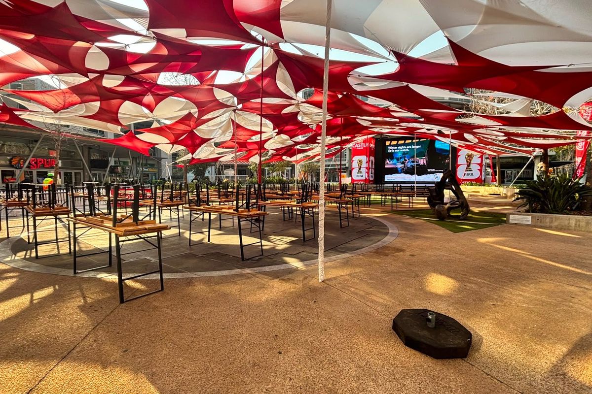 A spacious outdoor entertainment area featuring a large tension fabric canopy, paved flooring, and a prominent digital display screen for public viewing.