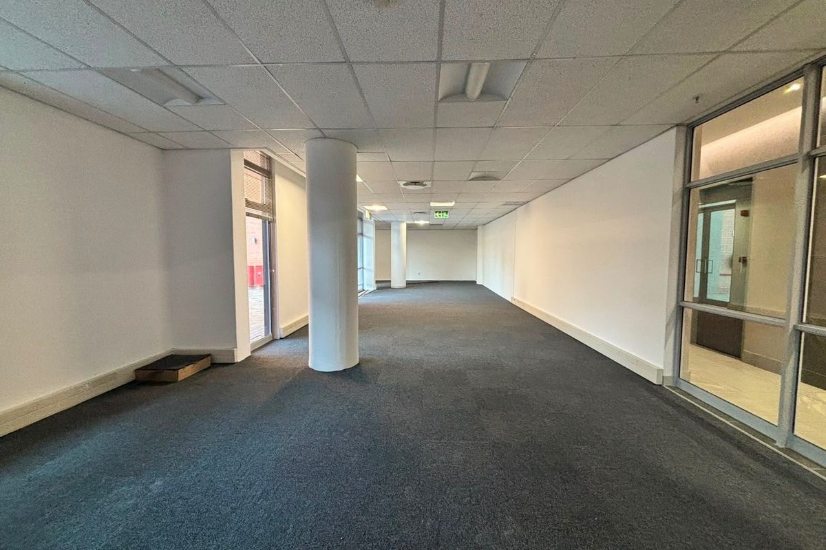 Spacious open-plan commercial office area featuring a suspended ceiling, structural columns, carpeted flooring, and large glass partitions providing natural light.