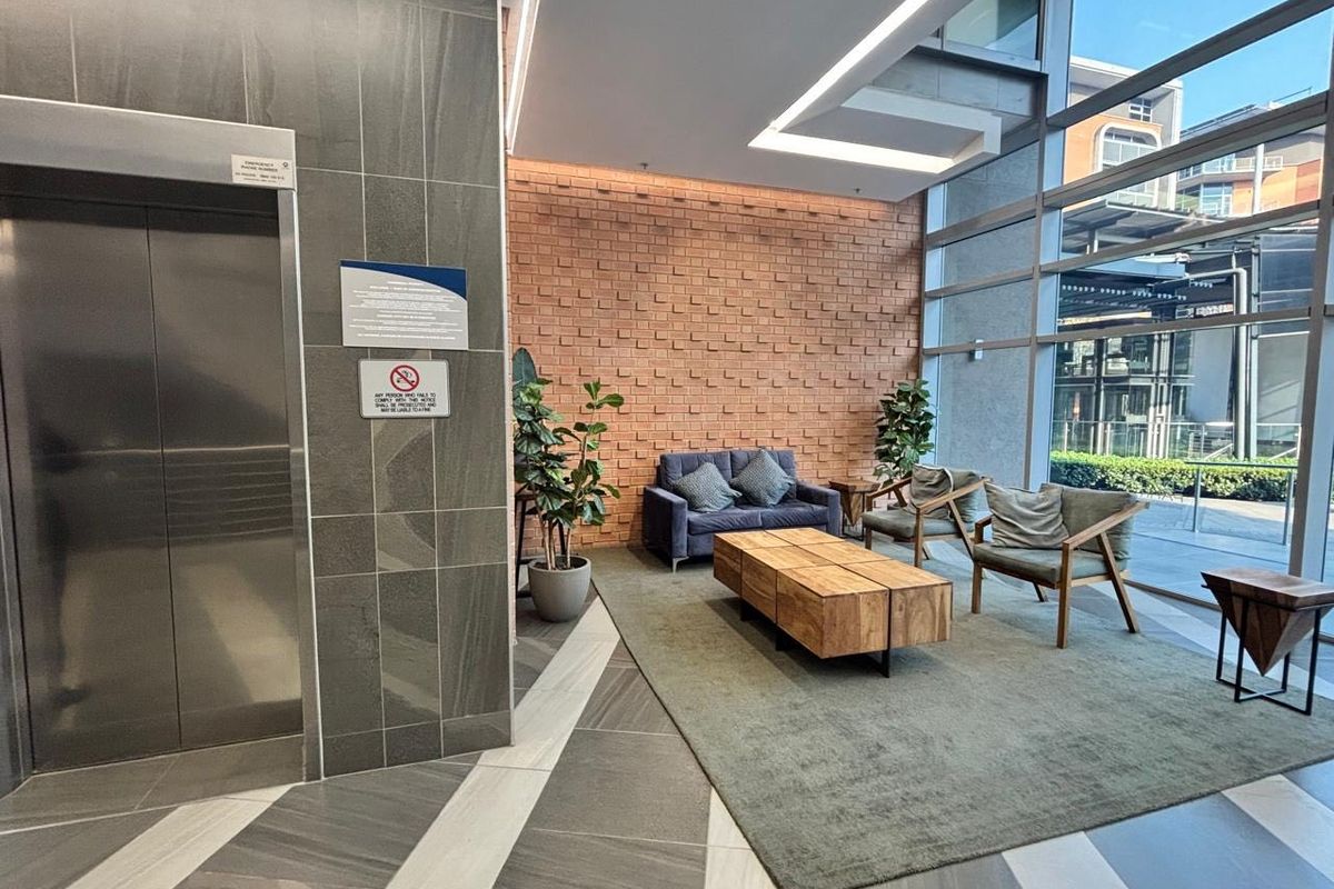 Modern commercial building lobby featuring a stainless steel elevator, exposed brick accent wall, and floor-to-ceiling glass windows providing ample natural light.