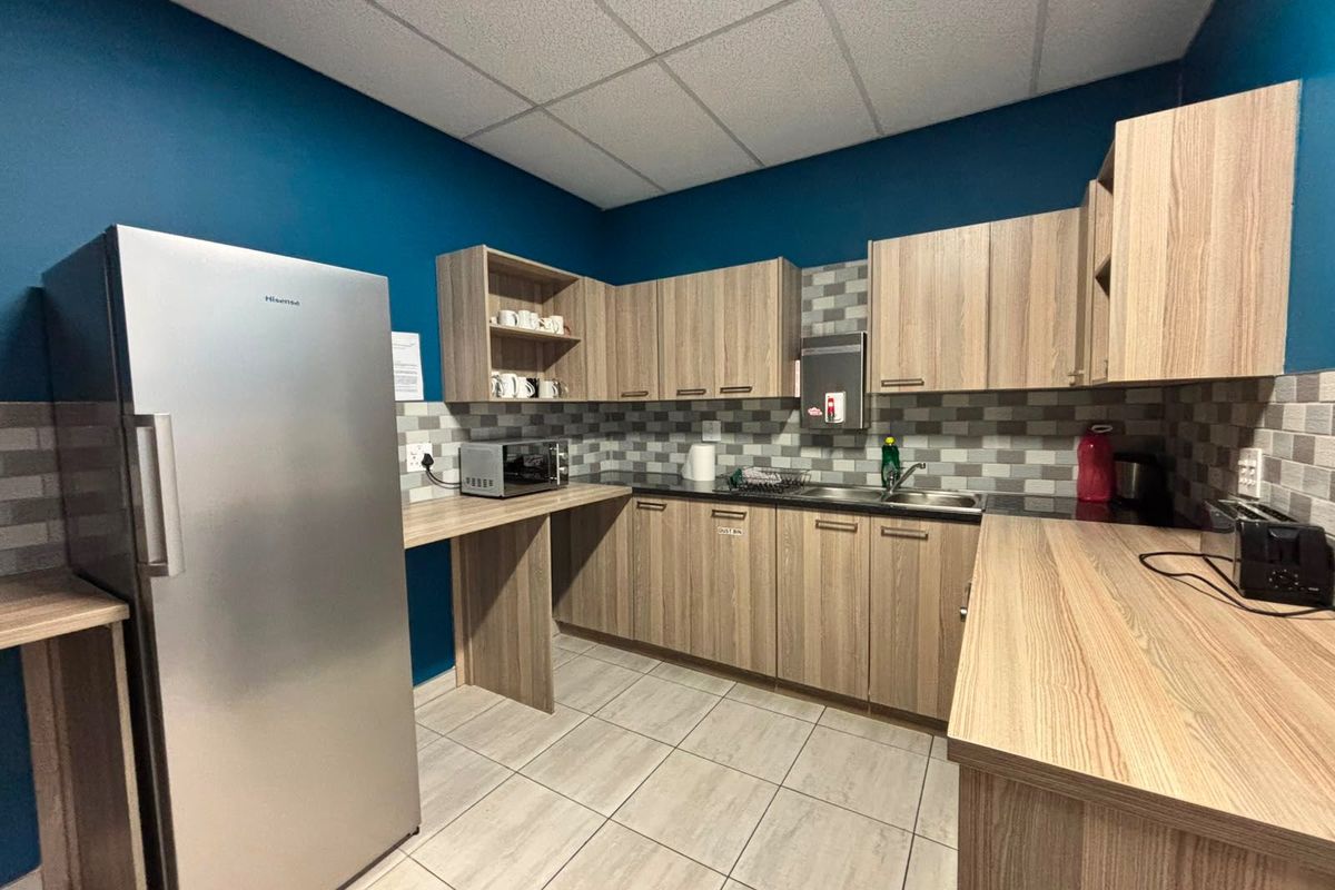 A functional commercial kitchen area featuring wood-look laminate cabinetry, tiled backsplash, stainless steel sink, and durable tiled flooring.