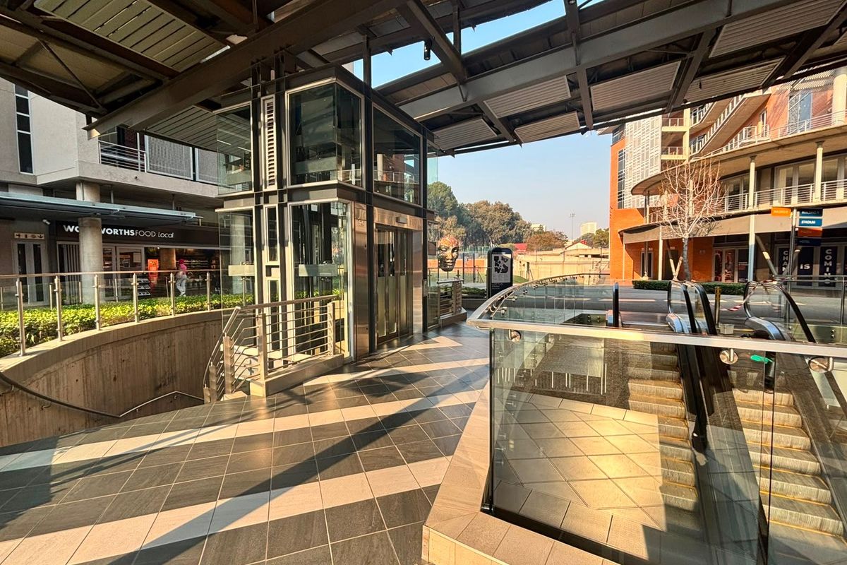Modern commercial exterior plaza featuring a glass elevator, escalator access, and high-quality tiled flooring under a structured metal canopy.