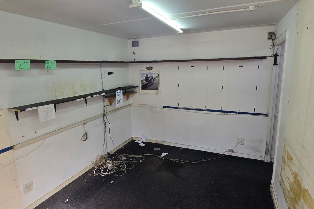 A vacant utility or storage space featuring wall-mounted shelving, fluorescent lighting, and electrical infrastructure, requiring cosmetic refurbishment and wall repairs.