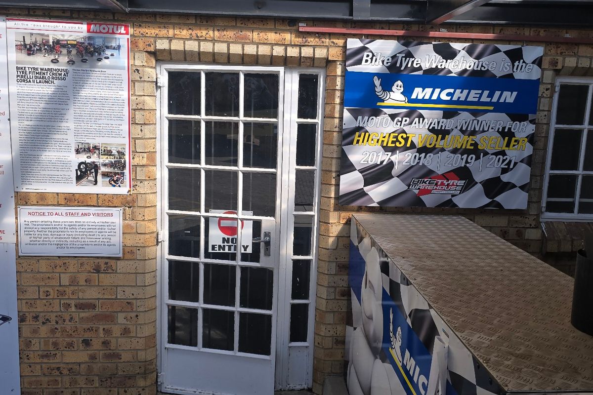 Industrial entrance area featuring face brick exterior, multi-pane glass door, and windows, with various commercial signage displayed on the facade.