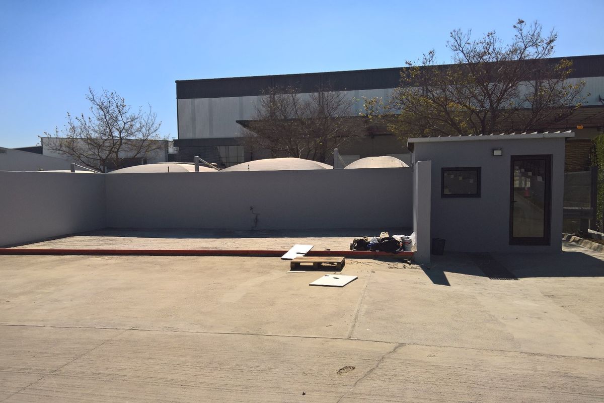 An industrial exterior area featuring a concrete yard, a perimeter wall, and a small security office building with a glass entrance door.