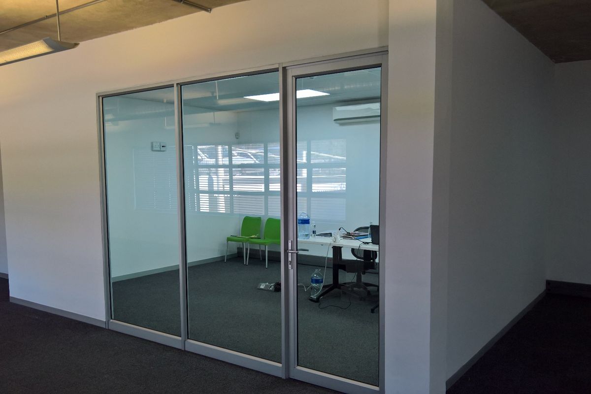 A modern office space featuring glass partitioning, carpeted flooring, and an exposed concrete ceiling with integrated lighting and air conditioning.