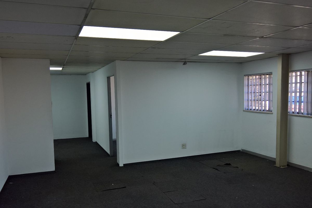 An empty industrial office space featuring a suspended ceiling, recessed lighting, security bars on windows, and worn carpet flooring requiring attention.