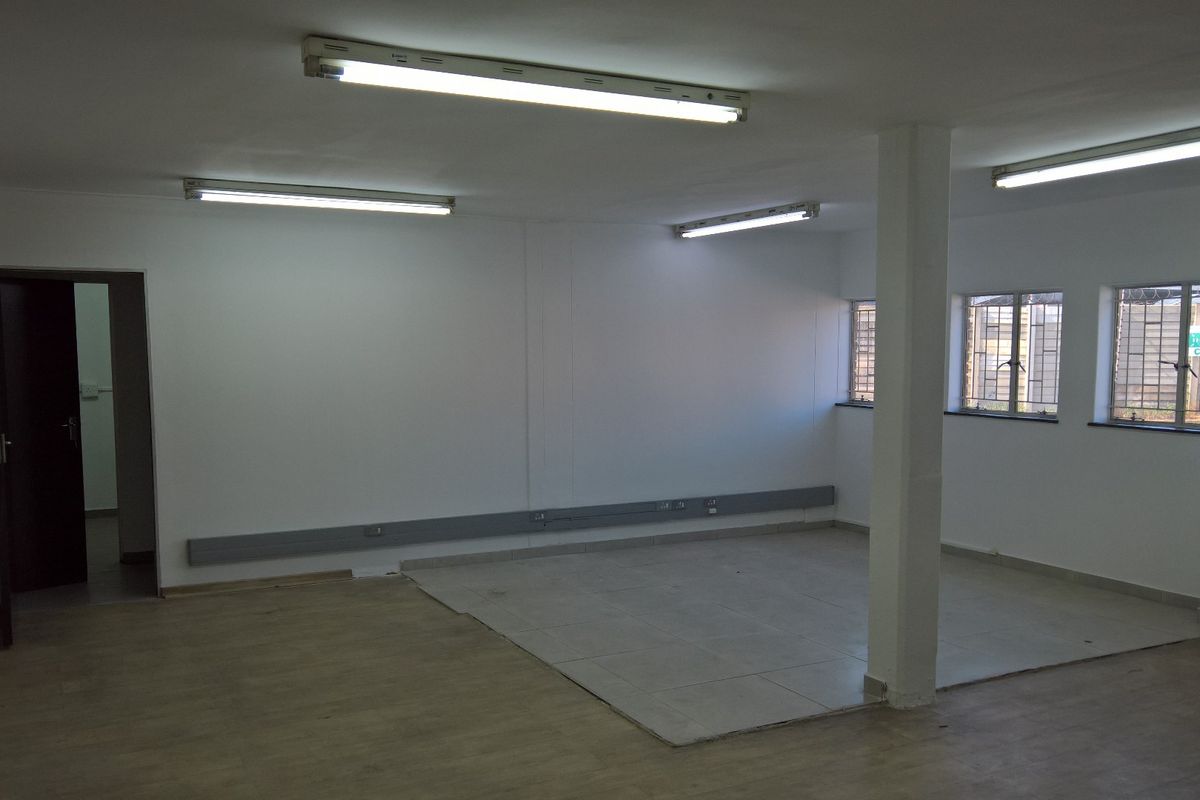 An open-plan industrial office space featuring a mix of tiled and laminate flooring, fluorescent lighting, and security-barred windows.