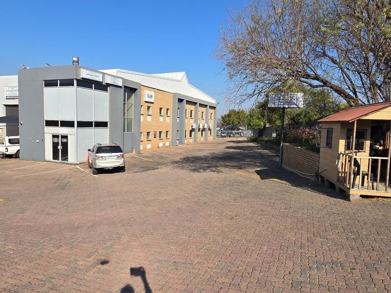 Front exterior view of an industrial warehouse complex featuring a paved driveway, multiple roller shutter doors, and a dedicated security guard hut.