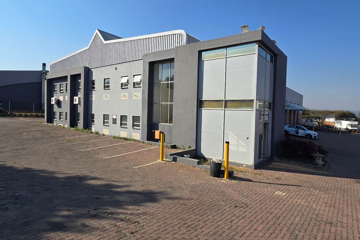 Front exterior view of a modern industrial building featuring a paved parking area, metal cladding, and a prominent glass facade section.