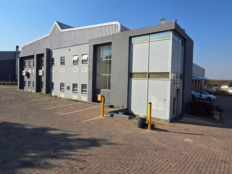 Front exterior view of a modern industrial building featuring a paved parking area, metal cladding, and a prominent glass facade section.