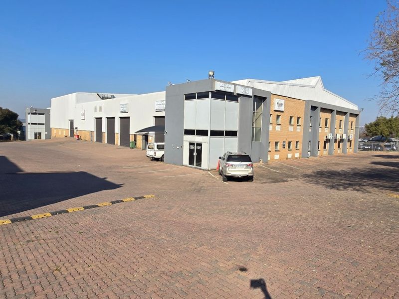 Exterior view of a large industrial warehouse facility featuring a paved parking area, multiple roller shutter doors, and a multi-level office section.