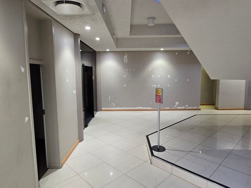 Spacious industrial building entrance lobby featuring tiled flooring, a central staircase with metal railings, and elevator access, currently requiring cosmetic wall repairs.