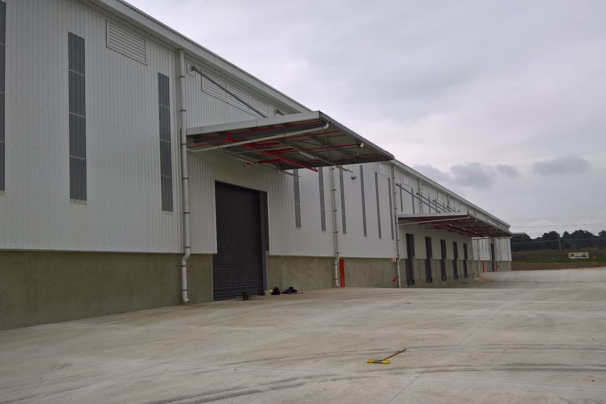 Modern industrial warehouse exterior featuring corrugated metal cladding, multiple roller shutter loading doors, and extensive concrete hardstand area.