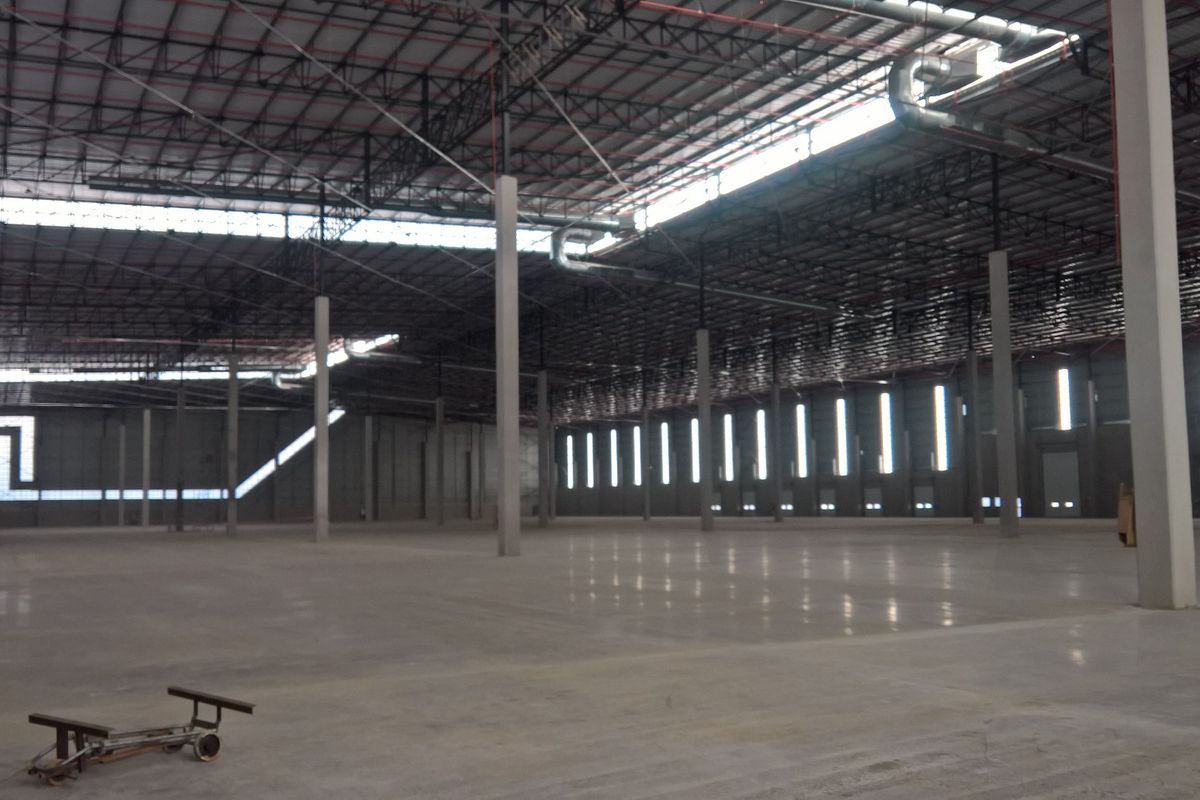 A vast, open-plan industrial warehouse space featuring polished concrete floors, high-clearance steel truss roofing, and extensive natural lighting through clerestory windows.