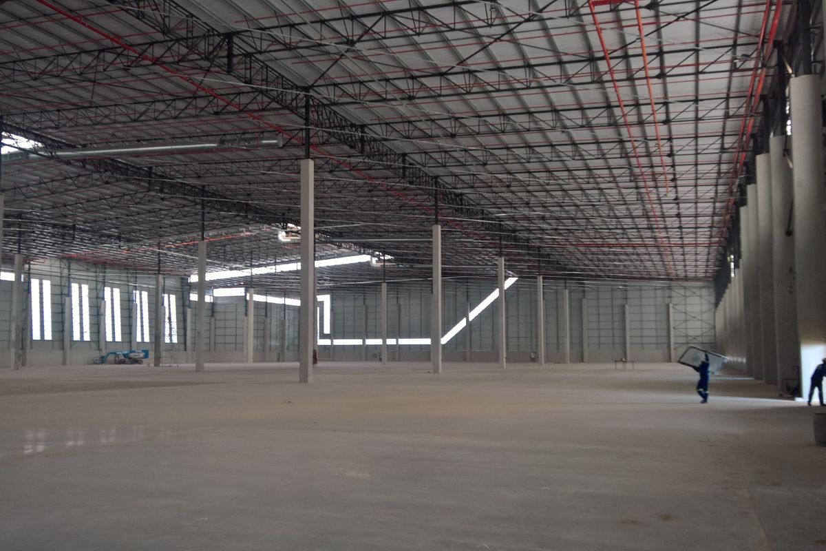 A large, open-plan industrial warehouse space featuring high ceilings with exposed steel trusses, fire sprinkler systems, and polished concrete flooring.