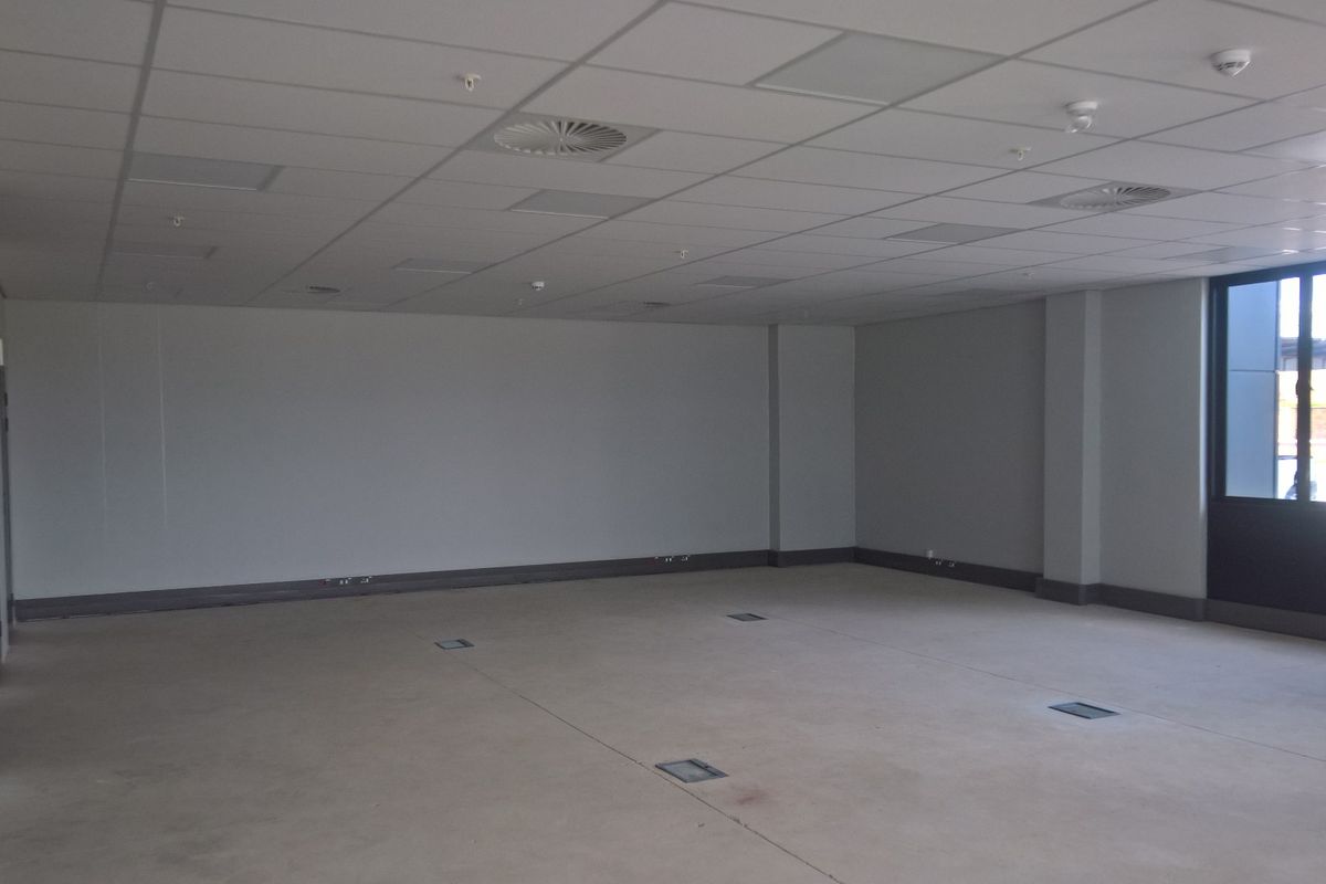 A spacious, vacant industrial office area featuring a suspended ceiling, recessed lighting, floor power access points, and large windows providing natural light.