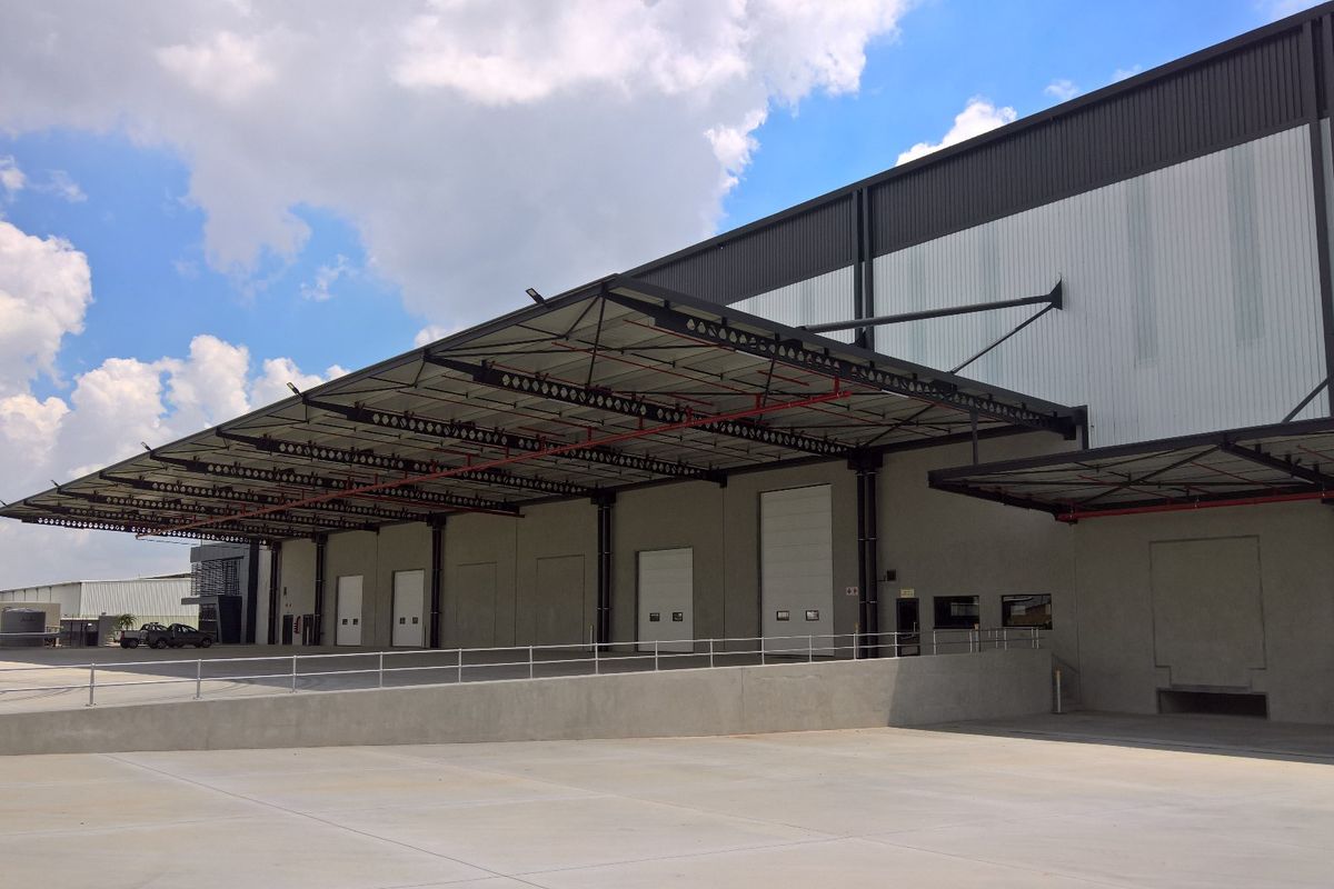 Modern industrial warehouse exterior featuring multiple roller shutter doors, a large concrete loading apron, and an extensive steel canopy structure.