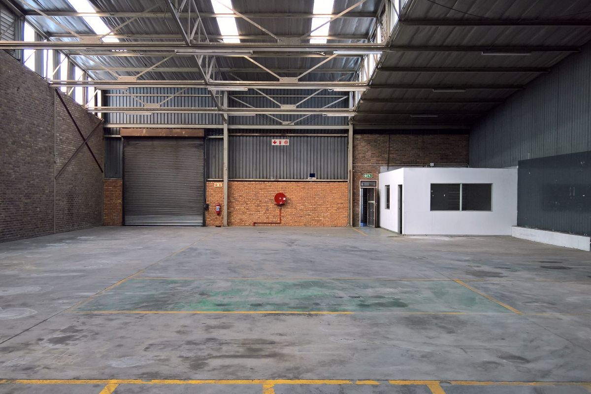 Spacious industrial warehouse featuring high ceilings with exposed steel trusses, a large roller shutter door, concrete flooring, and an integrated internal office structure.