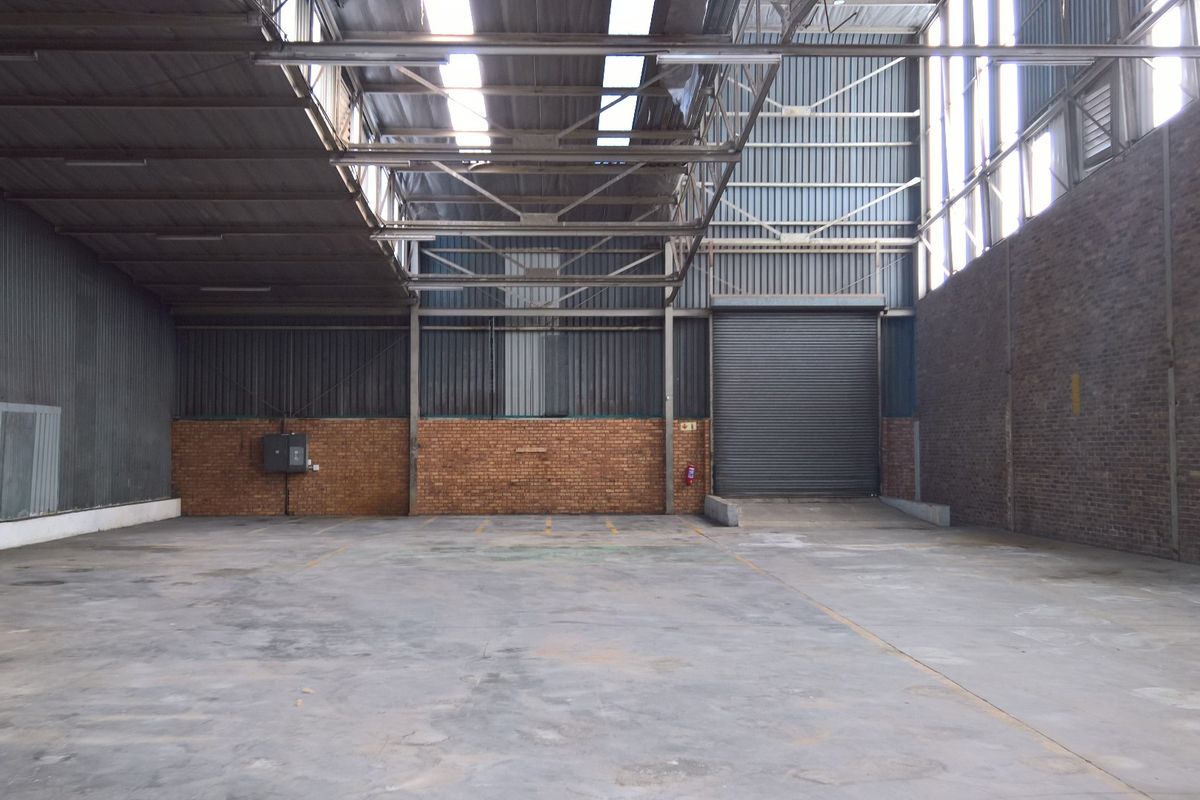 Spacious industrial warehouse unit featuring high ceilings with exposed steel trusses, concrete flooring, brick walls, and a large roller shutter door for easy access.