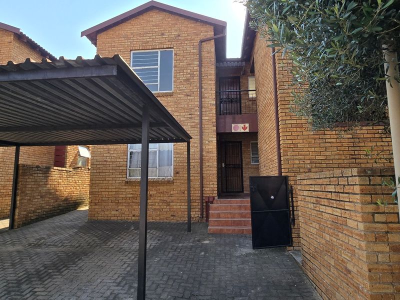 Front exterior view of a facebrick townhouse featuring a covered carport, paved driveway, and secure entrance with tiled steps.