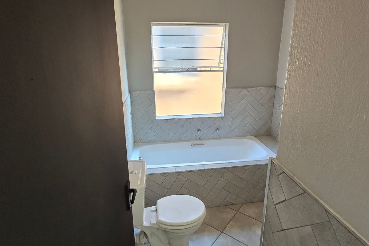 A clean bathroom featuring a built-in bathtub with herringbone tile surround, a standard toilet, and tiled flooring with natural light from a louvered window.