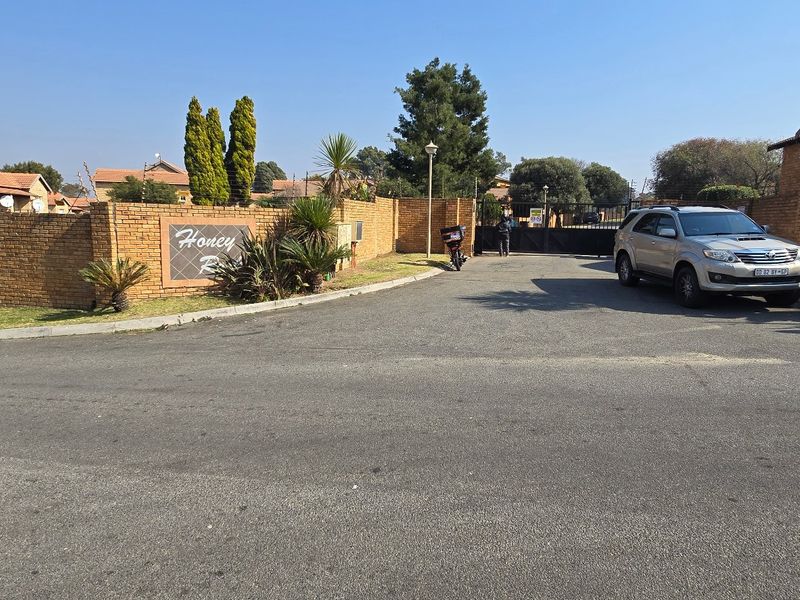 Street view of the townhouse complex entrance featuring a brick boundary wall, security gate, and paved access road.
