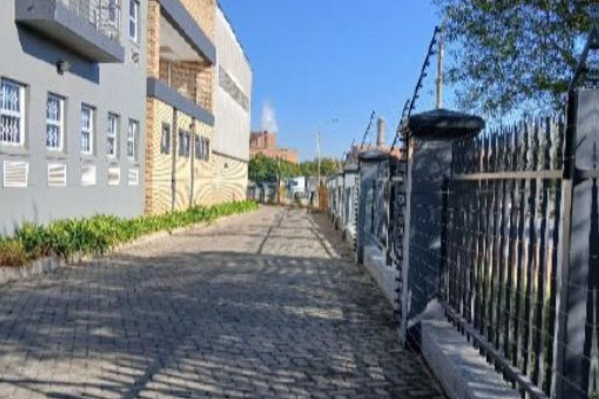 Exterior view of an industrial property featuring a cobblestone driveway, secure perimeter fencing with electric wires, and a multi-story building with balconies.