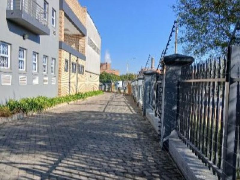 Exterior view of an industrial property featuring a cobblestone driveway, secure perimeter fencing with electric wires, and a multi-story building with balconies.