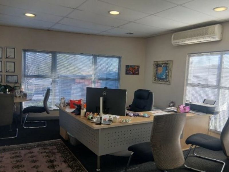 A functional office space featuring suspended ceiling tiles, recessed lighting, and a wall-mounted air conditioning unit with large windows and venetian blinds.